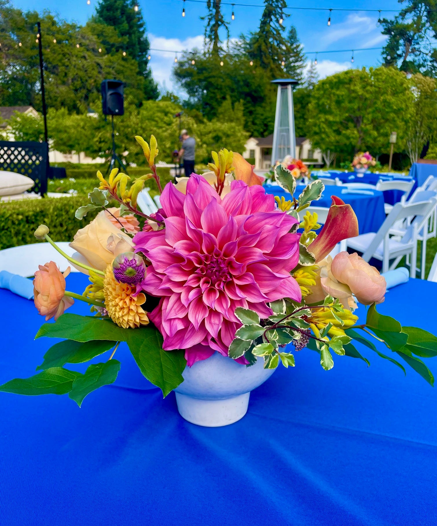 Floral centerpiece arrangement with a large pink dahlia in a modern white vase on a blue tablecloth outdoors.